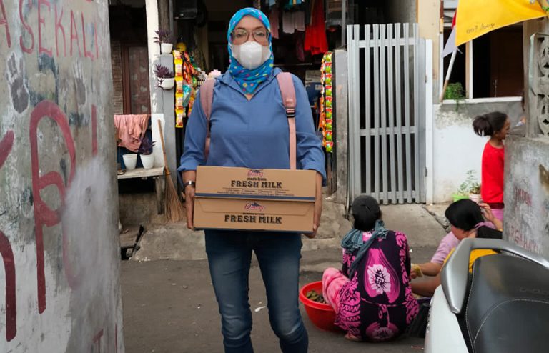 Seorang perempuan menggunakan jaket tudung dan masker membawa satu kardus susu dalam kemasan. Perempuan penjangkau untuk anak yang hidup dengan HIV di Indonesia oleh Lentera Anak Pelangi.